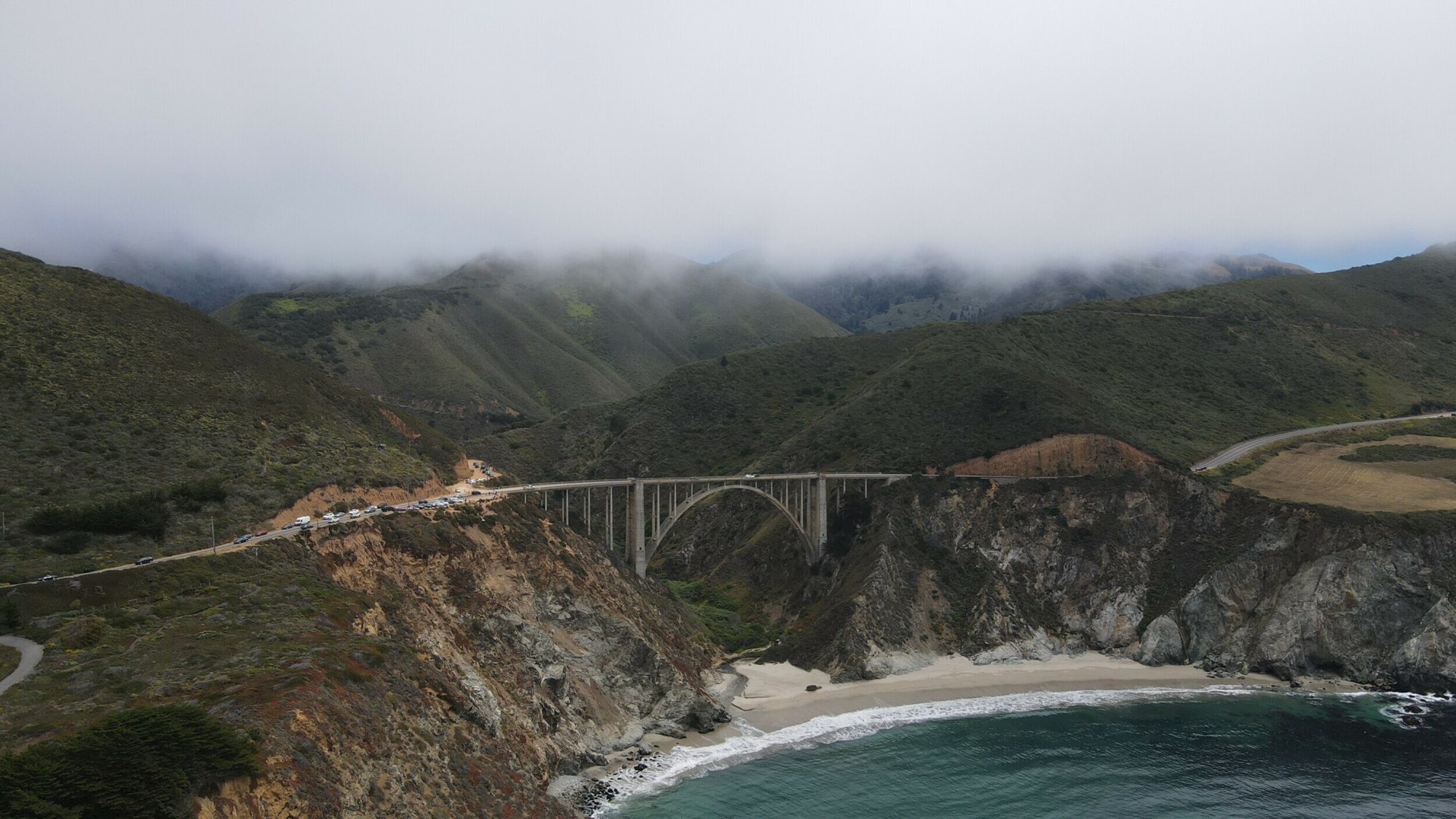 Aerial photograph of Bixby Bridge enveloped in fog, with the Pacific Ocean coastline and green mountains.