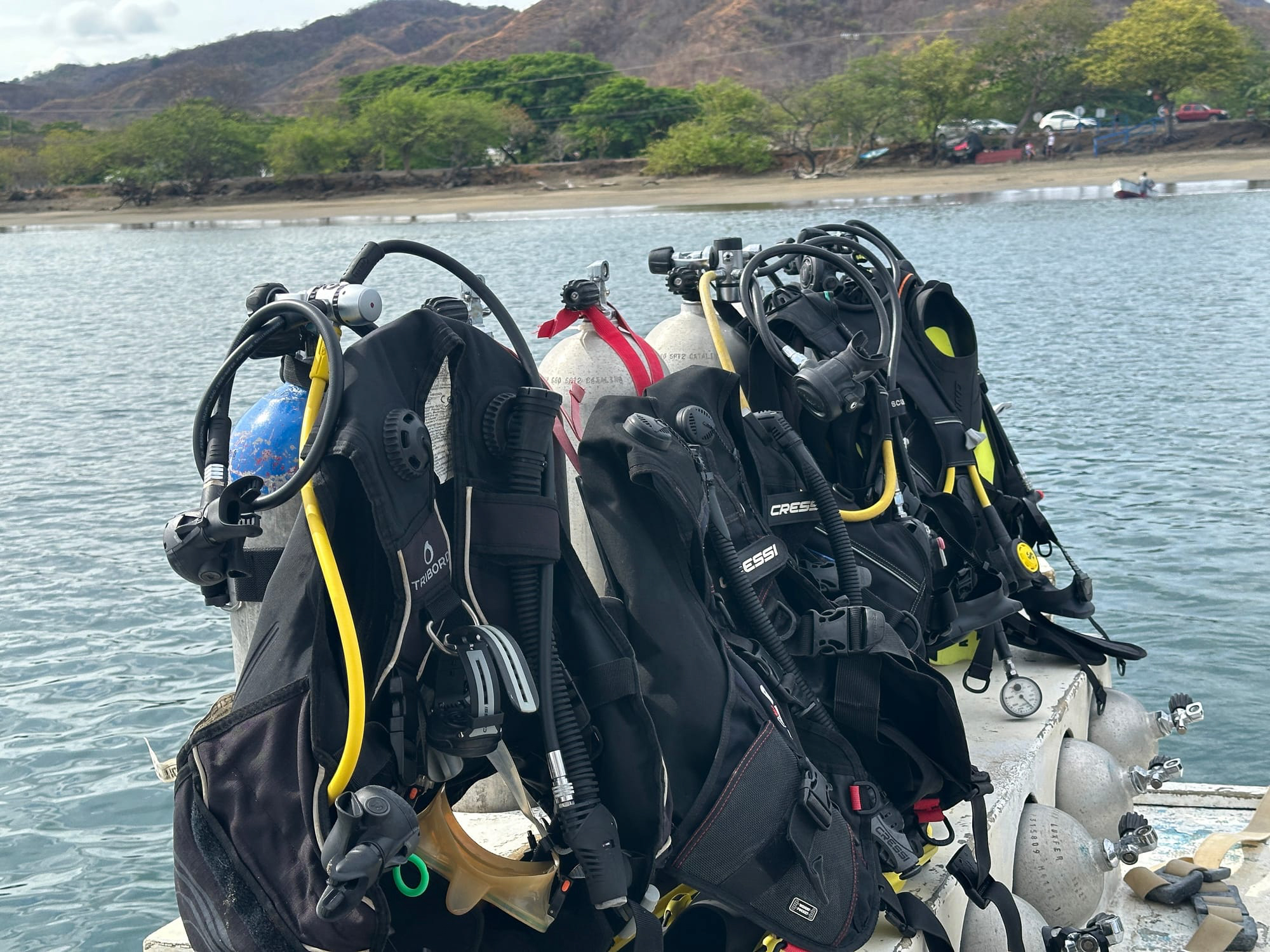 Scuba diving gear lined up on a dock near the coast of Tamarindo, Costa Rica, with hills and a beach in the background.