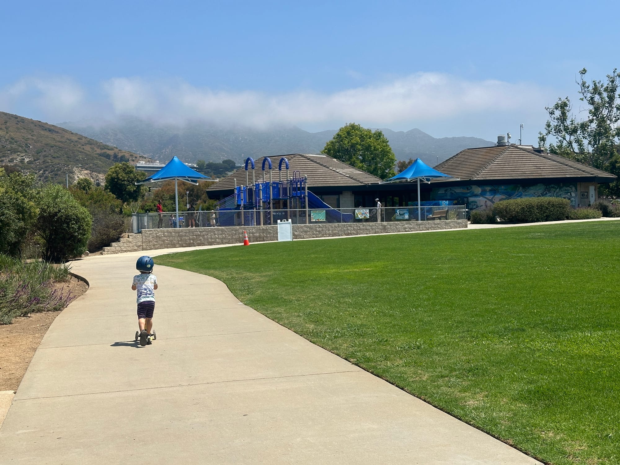  A child rides a scooter at Malibu Bluffs Park, with a playground and hills in the background.