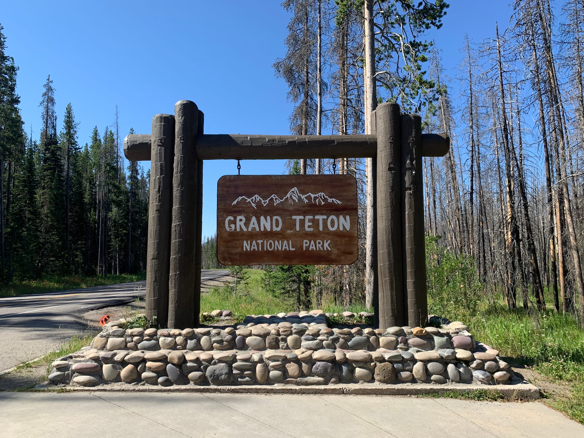 A wooden sign marking the entrance to Grand Teton National Park, surrounded by tall trees and a clear blue sky