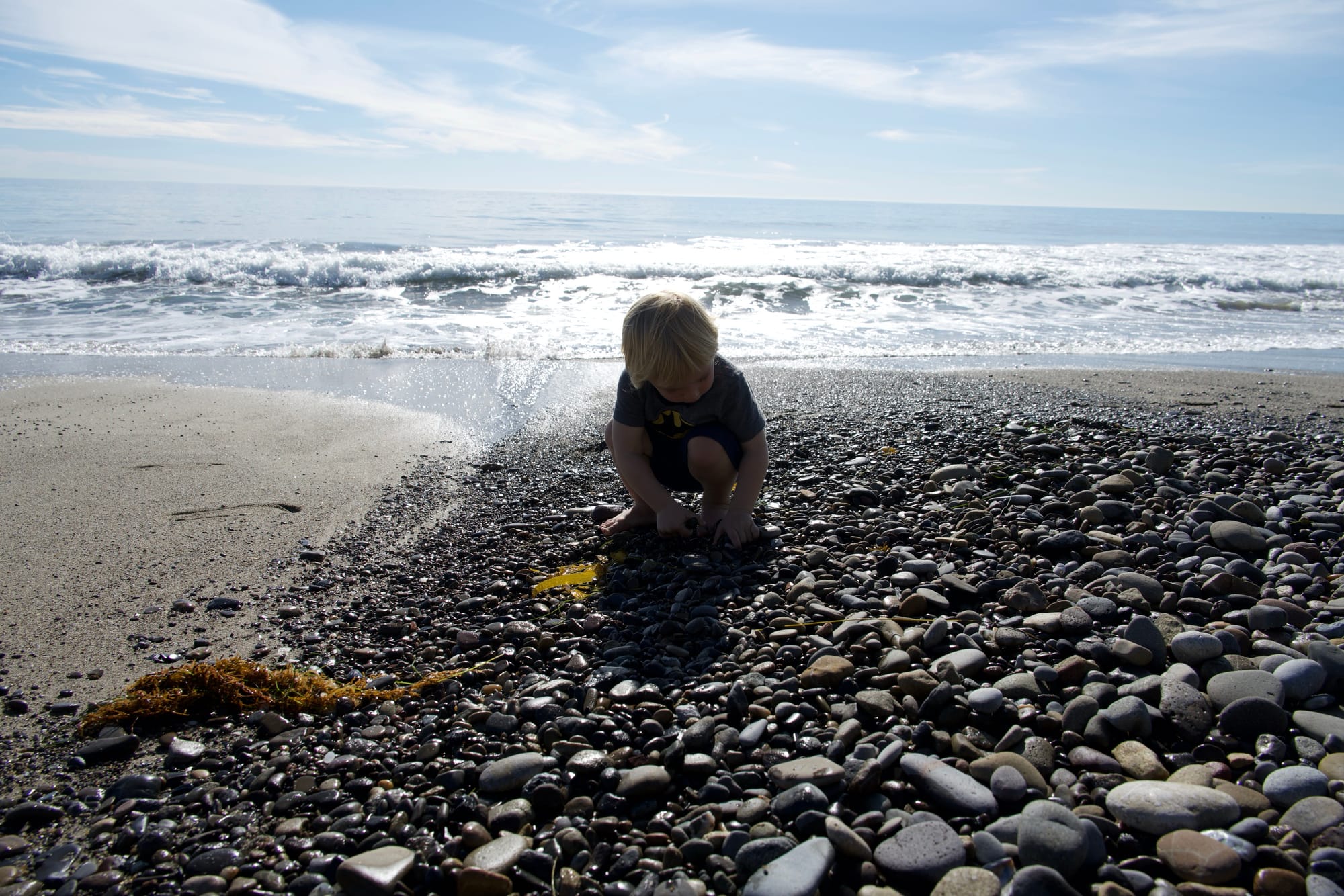 A young child crouching on a rocky Malibu beach with ocean waves in the background.