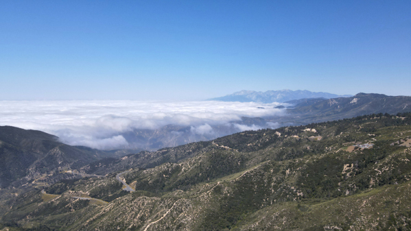 Aerial view of Big Bear, California's mountains with clouds below and clear blue sky above.