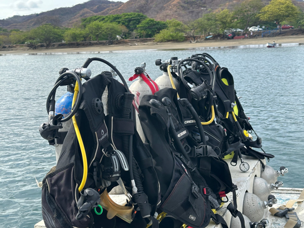 Scuba diving gear lined up on a dock near the coast of Tamarindo, Costa Rica, with hills and a beach in the background.