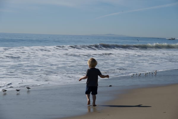 A young child stands barefoot on the beach, facing the ocean with outstretched arms.