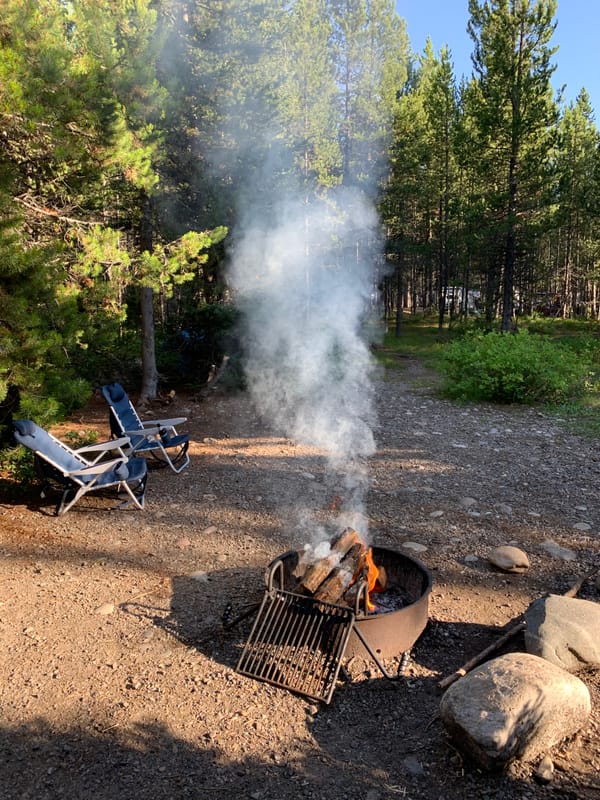 Logs burning with smoke rising from a campfire at a peaceful campsite surrounded by trees in Grand Teton National Park.