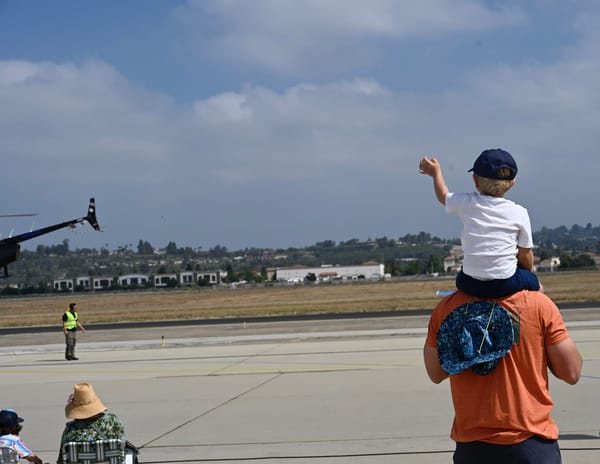 A child sitting on his father's shoulders waves at an aircraft on the runway during the Wings Over Camarillo Airshow.