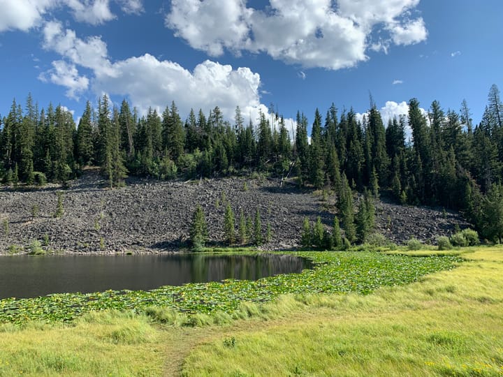 A pond surrounded by meadows, with a hillside and rocky terrain in the background under the sky in Yellowstone NP.
